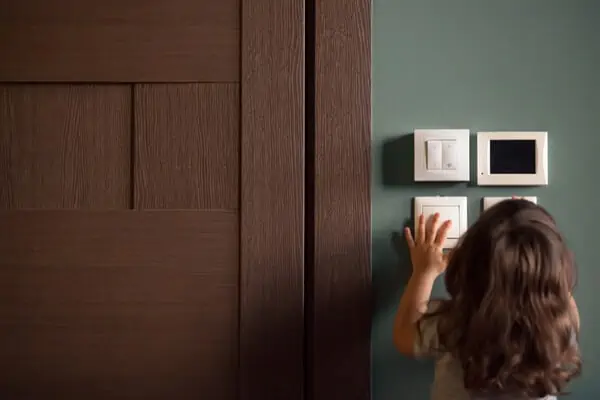 Brun dörr. intill står ett barn med långt brunt hår och justerar strömbrytare på vägg.